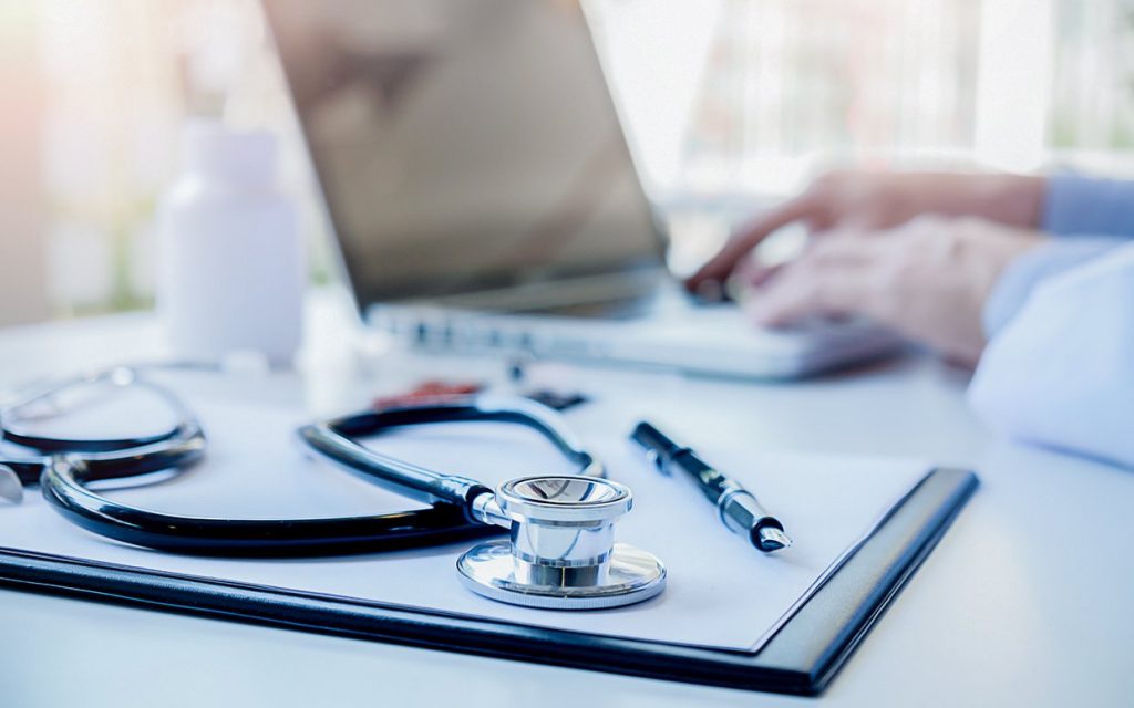 Médico digitando no computador sob mesa com estetoscópio caneta