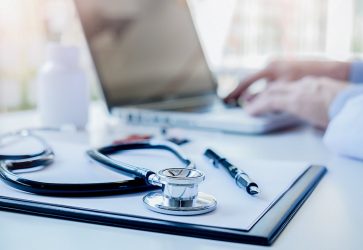 Médico digitando no computador sob mesa com estetoscópio caneta