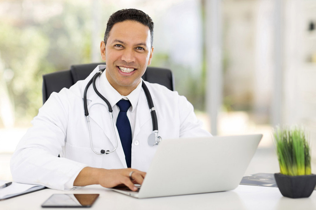 Médico sorrindo sentado digitando no computador | O que é o e-Médico e como ele pode ser utilizado?